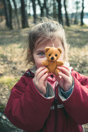 Little smilling happy girl playing with her little teddy bear toy in a park on sunny spring day. Child holding teddy at front of face looking at camera wearing red jacketの写真素材