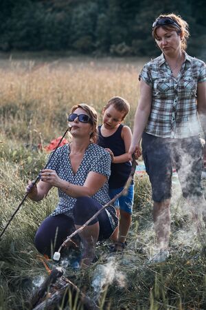 Women roasting a marshmallows over a campfire on meadow. Vacations close to nature. Candid people, real moments, authentic situationsの写真素材