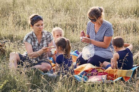 Families and friends spending time together on a meadow, close to nature. Mothers feeding kids, sitting on a blanket, on grass. Candid people, real moments, authentic situationsの写真素材