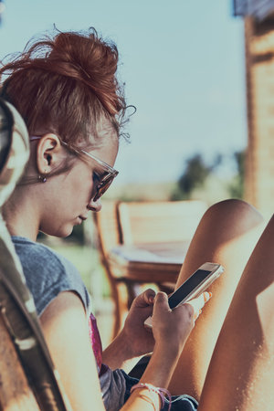 Young woman talking on a  mobile phone sitting in a chair outdoors on patio wearing a sunglasses. Real people, authentic situationsの写真素材