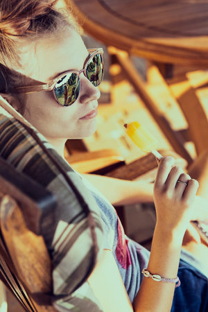 Young woman eating a ice cream, sitting in a chair outdoors on a patio during summer day. Real...