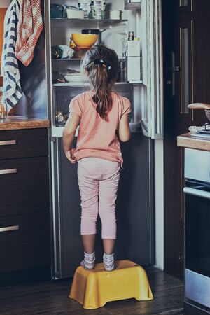 Little girl standing on a childs stool in front of opened fridge, looking for ice creams among food products. Real people, authentic situationsの写真素材