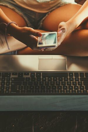 Young woman using portable computer and mobile phone, sitting on a bed, learning online at home. Candid people, real moments, authentic situationsの写真素材