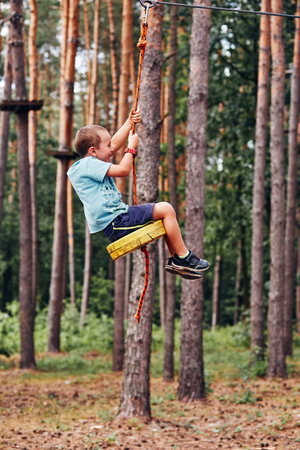 Happy boy riding on the zip line in rope park in forest while spending summer vacation. Real people,...