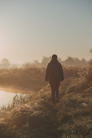 Woman walking through a meadow by a pond in the foggy morning. Sun rising above field and pond flooded with fog in the morning. Real people, authentic situationsの写真素材
