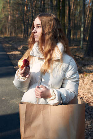 Young woman carrying big paper shopping bag. Girl standing in a park and looking the side, holding apple, wearing white jacket. Real people, authentic situationsの写真素材
