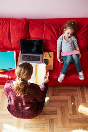 Woman mother working doing her job remotely during video chat call stream online course webinar on laptop from home during COVID-19 quarantine while her daughter watching video on tablet. Woman sitting on bed in front of computer looking at screenの写真素材