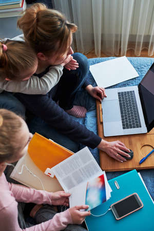 Women mother and daughter working learning remotely doing job homework during video chat call stream online course webinar lessons on laptop from home during COVID-19 quarantine. Women sitting on bed in front of computer looking at screen in company little girl playing aroundの写真素材