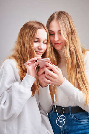 Young women girls listening to music together streaming content having fun watching video enjoying video chat talking with friends making gestures faces using smartphone earphones headphones standing over plain grey backgroundの写真素材