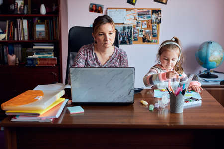 Woman mother working doing her job remotely during video chat call stream online course webinar on laptop from home while her daughter making drawings playing around and listening to stories. Woman sitting at desk in front of computer looking at screenの写真素材
