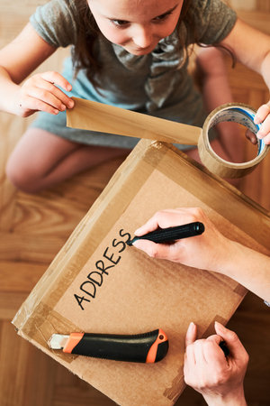 Woman writing an address on a cardboard box parcel in room at home. Little girl helping her mother...