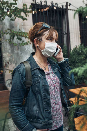 Young woman talking on phone standing in a street downtown. Woman is wearing the face mask to avoid virus infection and to prevent the spread of disease in time of coronavirusの写真素材