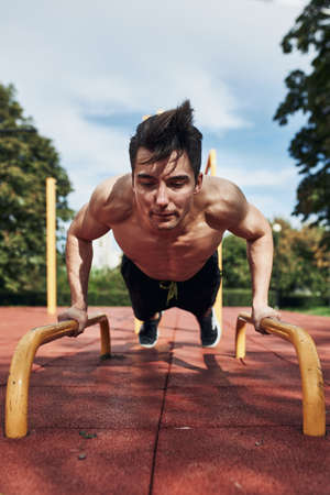 Young shirtless man bodybuilder doing push-ups on a parallel bars during his workout in a modern calisthenics street workout parkの写真素材
