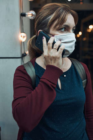 Young woman talking on phone standing in a front of restaurant downtown wearing the face mask to avoid virus infection and to prevent the spread of disease in time of coronavirusの写真素材