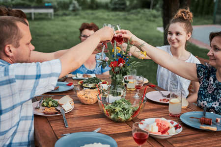 Family making toast during summer picnic outdoor dinner in a home garden. Close up of people holding wine glasses with red wine over table with food and dishesの写真素材