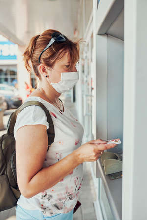 Woman withdrawing money from atm machine using debit or credit card standing outdoors in the street, wearing face mask to cover mouth and nose during pandemic coronavirus outbreakの写真素材