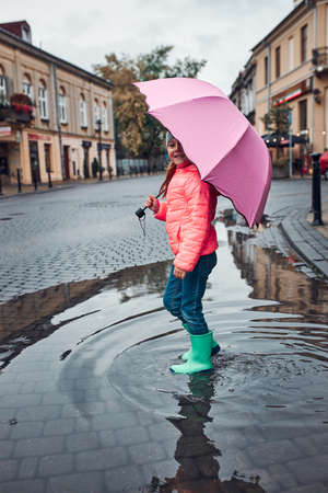 Child holding big pink umbrella walking through the puddle in a downtown on rainy gloomy autumn dayの写真素材