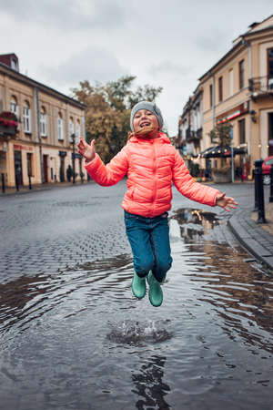 Happy smiling girl jumping in the puddle during walk in a downtown on rainy gloomy autumn dayの写真素材