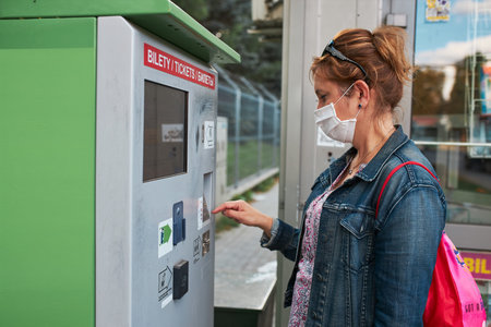 Woman buying bus ticket in ticket machine wearing face mask to cover mouth and nose traveling during...