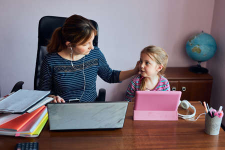 Woman mother working doing her job remotely during video chat call stream online course webinar on laptop from home while her daughter watching video on tablet. Woman sitting at desk in front of computer looking at screenの写真素材