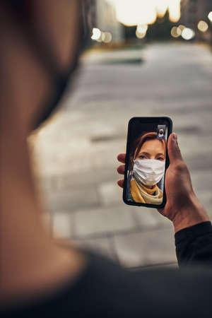 Young man having video call with her mother standing outdoors in the street in the evening wearing the face mask to avoid virus infection and to prevent the spread of disease in time of coronavirusの写真素材