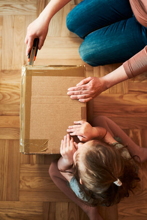 Woman unpacking a cardboard box parcel in room at home. Little girl waiting for opening a gift in...