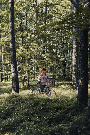 Active woman spending free summer vacation time on a bicycle trip in a forest. Woman wearing bicycle helmet and gloves holding bike with basketの写真素材
