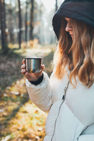 Woman in a hood having break during autumn trip holding cup with hot drink from thermos flask on autumn cold day. Active girl wandering in a forest actively spending timeの写真素材