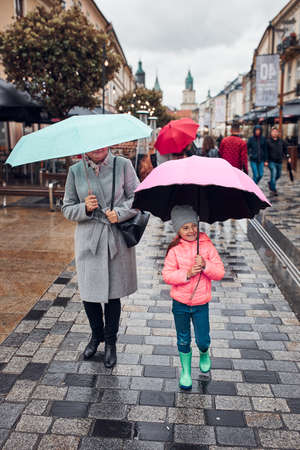 Mother and her little daughter holding the pink and blue umbrellas walking in a downtown on rainy gloomy autumn dayの写真素材