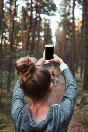 Young woman taking photos using smartphone during walk in a forest on summer vacation trip. Back view of girl holding smartphoneの写真素材