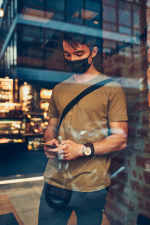 Young man standing in coffee shop at store front in the city center in the evening, wearing the face...