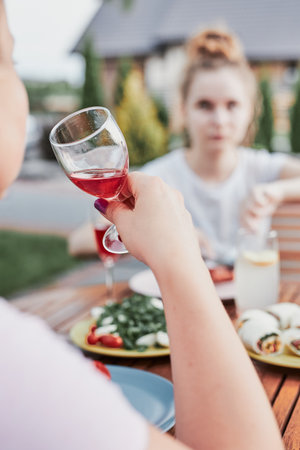 Woman drinking red wine during summer outdoor dinner in a home garden. Close up of hand holding wine glass with red wine over table with dishesの写真素材