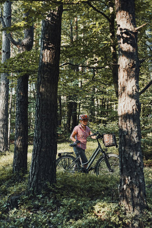 Active woman spending free summer vacation time on a bicycle trip in a forest. Woman wearing bicycle helmet and gloves holding bike with basket standing behind the treesの写真素材