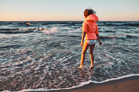 Playful little girl jumping over sea waves on a sand beach at sunset during summer vacationの写真素材