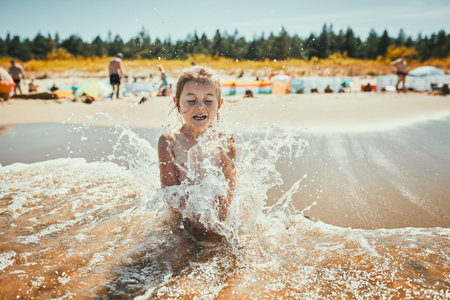 Little girl splashed by sea wave while sitting and playing on a beach. Child enjoying the sea and sunlight during summer vacationの写真素材