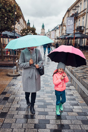 Mother and her little daughter holding the pink and blue umbrellas walking in a downtown on rainy gloomy autumn dayの写真素材