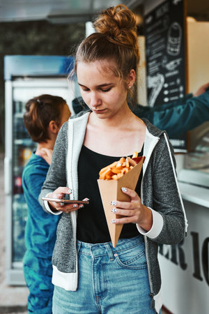 Teenage girl eating potato fries from carton cone standing in front of food truck. Teenager having a fast food meal outdoors during summer vacationsの写真素材
