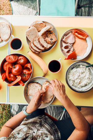 Breakfast prepared during summer vacation on camping. Bread, cottage cheese, cold meat, tomatoes, fruits and coffee cups on table. Close up of outdoor table setting set under tentの写真素材