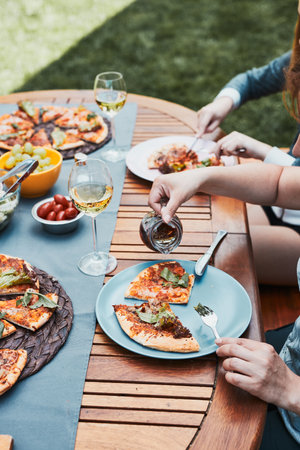 Family and friends having meal - pizza, salads, fruits and drinking white wine during summer picnic outdoor dinner in a home garden. Close up of people sitting at the table in a orchard in a backyardの写真素材