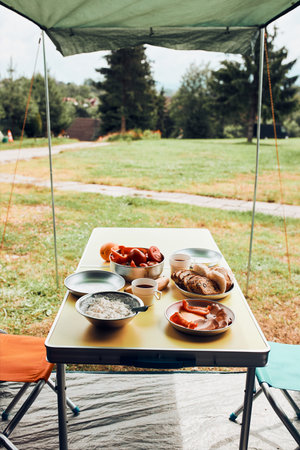 Breakfast prepared during summer vacation on camping. Bread, cottage cheese, cold meat, tomatoes, fruits and coffee cups on table. Close up of outdoor table setting set under tentの写真素材