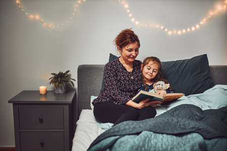 Mother reading book her daughter in bed before going to sleep. Reading stories before sleep. Bedtime stories for childの写真素材