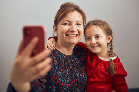 Mother with her little daughter making video call using mobile phone. Keeping distance. Woman and little girl talking with relatives. Cheerful family having fun taking selfie photo using smartphone. Connecting remotely with familyの写真素材