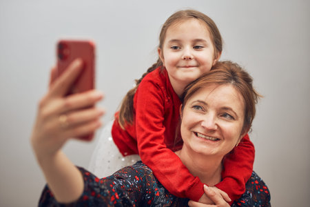 Mother with her little daughter making video call using mobile phone. Keeping distance. Woman and little girl talking with relatives. Cheerful family having fun taking selfie photo using smartphone. Connecting remotely with familyの写真素材