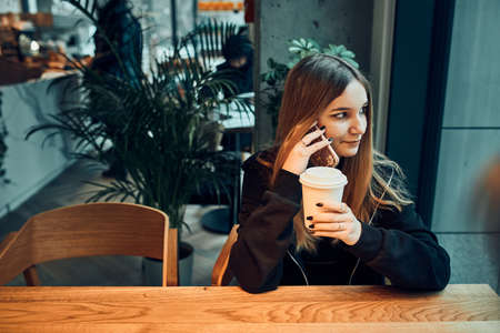 Young woman talking on the phone at cafe, having a pleasant phone call, answering call, chatting by mobile phone with friend while sitting in a cafe and drinking coffee. Girl relaxing in cafeの写真素材