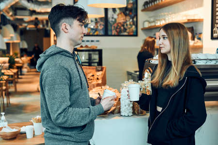 Friends having a chat, holding coffee cups, standing in a coffee shop and waiting for the orders. Man and woman having a break drinking coffee in cafeの写真素材