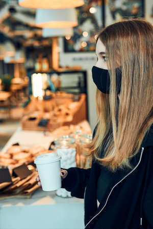Woman holding coffee cup, standing in queue in a coffee shop and waiting for the order. Person wearing the face mask to avoid virus infection and to prevent the spread of diseaseの写真素材