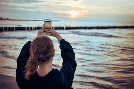 Young woman taking photos of sunset over sea using smartphone during summer trip. Rear view of girl holding smartphone and taking pictureの写真素材