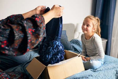 Mother and her daughter opening box with ordered dress at home on couch. Mom online shopper customer holding dress. Delivery service fashion conceptの写真素材