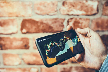 Person investing trading on stock cryptocurrency market using investing application on smartphone. Stock market investment in hand. Trader holding mobile phone looking at candle chartの写真素材