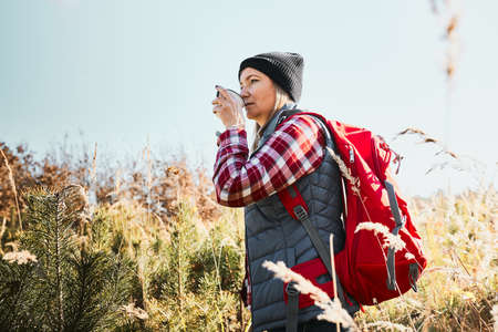 Woman taking break and relaxing with cup of coffee during summer trip. Woman standing on trail and looking away. Female with backpack hiking through tall grass along path in mountains. Spending summer vacation close to natureの写真素材